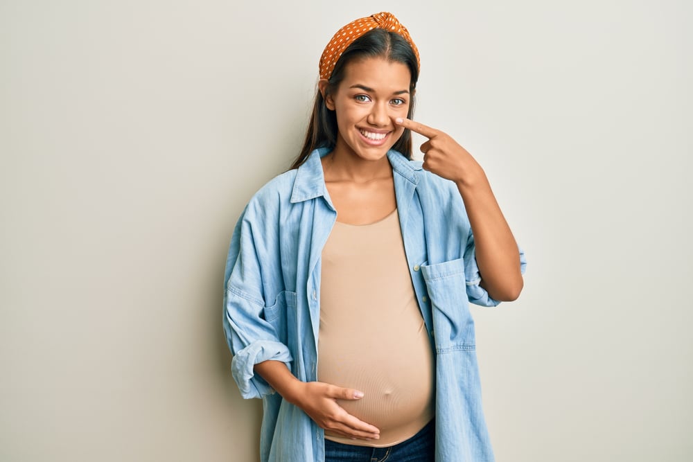 A pregnant rhinoplasty patient pointing to her nose, happy with the appearance of natural beauty obtained from their cosmetic treatment.