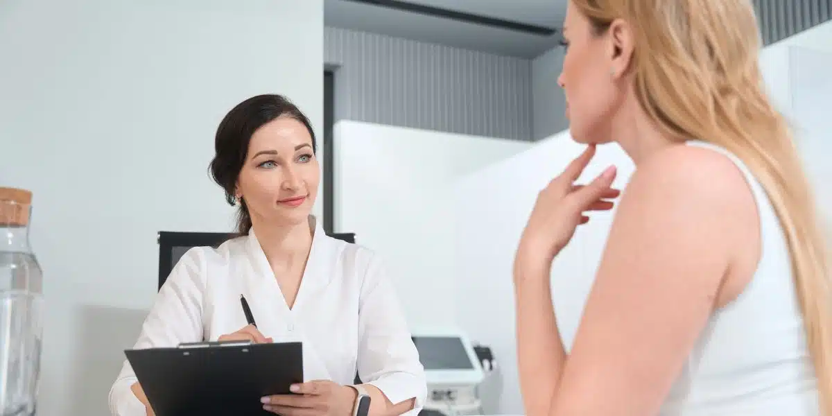 Female doctor holding a clipboard and discussing facelift costs with a blonde woman in a consultation room.