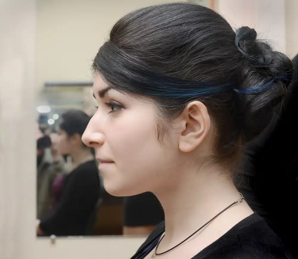 Side view of a woman with a hooked nose contemplating rhinoplasty in a mirror reflection.