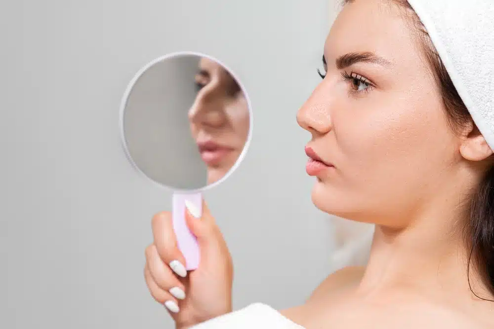 Woman in a bathrobe, with towel-wrapped hair, inspecting her nose in a handheld mirror post-rhinoplasty.