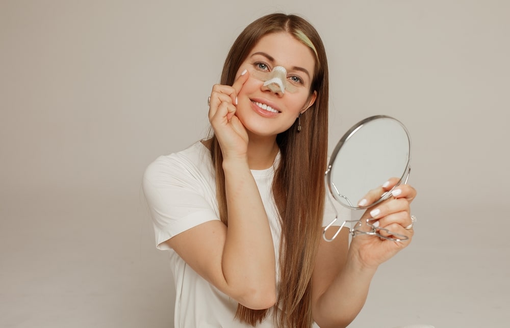 Woman with nasal dressing examining her post-operative nose in mirror.