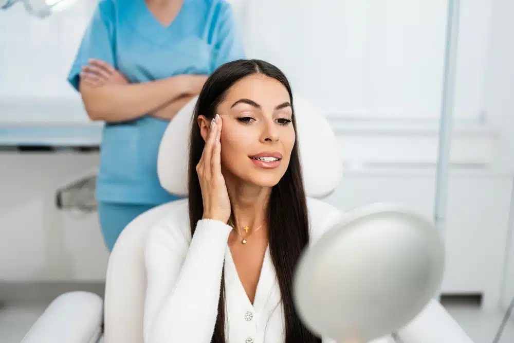 Woman with long dark hair looking at her face in a handheld mirror, sitting in a medical chair, with a medical professional standing in the background.