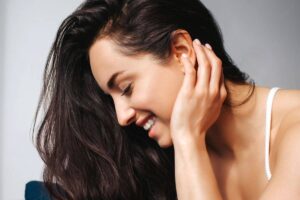 Woman smiling with head tilted and hand near ear, reflecting confidence and healing after otoplasty surgery.