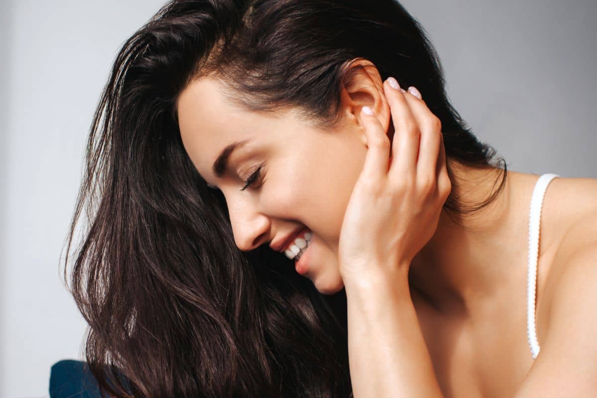 Woman smiling with head tilted and hand near ear, reflecting confidence and healing after otoplasty surgery.
