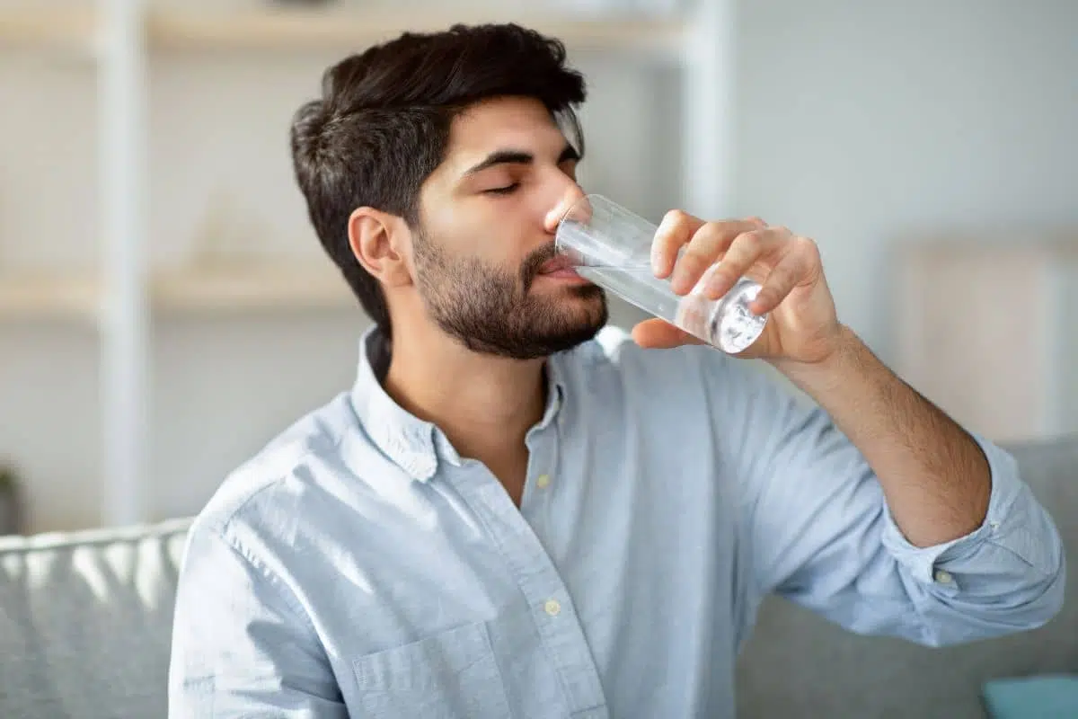 Man in casual shirt drinking water, illustrating healthy hydration as part of pre-rhinoplasty preparation steps.