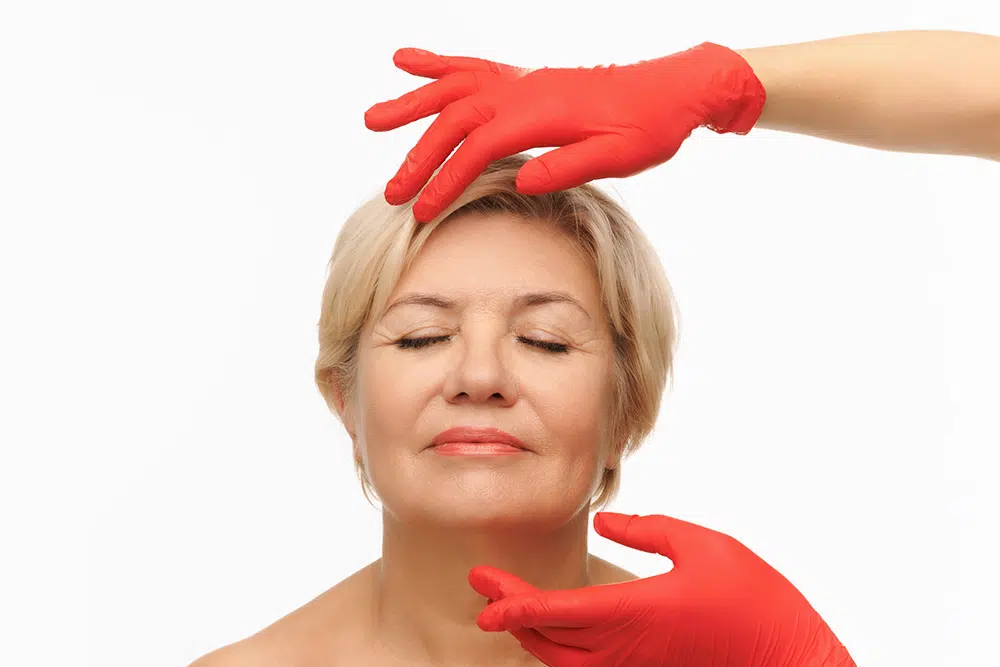 Mature woman with eyes closed receiving facial assessment before deep plane facelift for long-lasting rejuvenation.