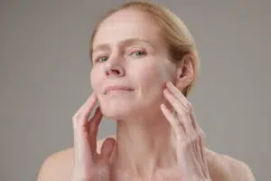 A woman gently touching her midface and cheek area to assess the firmness and degree of swelling following a deep plane facelift.