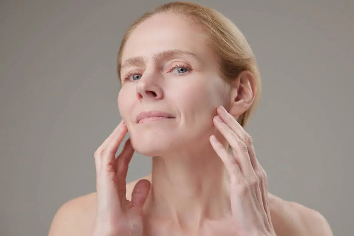 A woman gently touching her midface and cheek area to assess the firmness and degree of swelling following a deep plane facelift.
