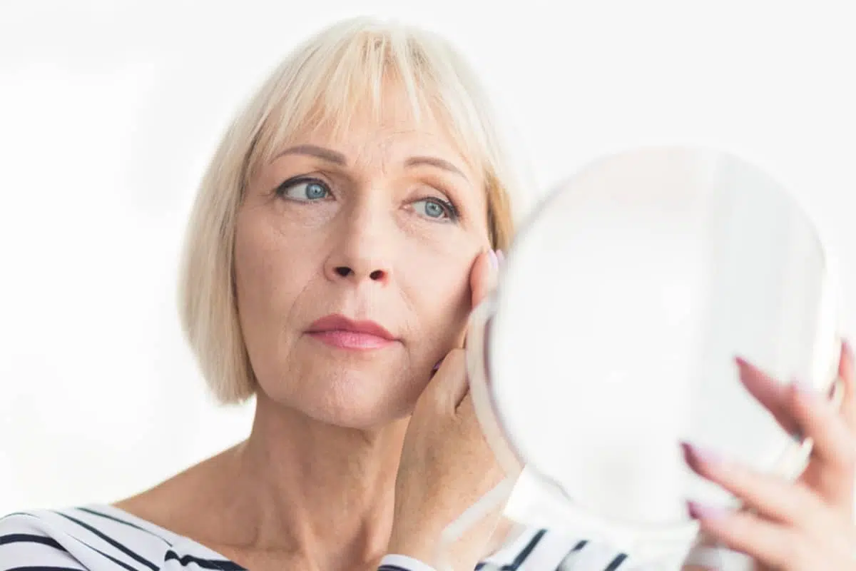 A woman with blonde hair looking into a handheld mirror to evaluate her facial contours and the gradual resolution of swelling after a facelift procedure.