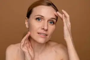 A woman touching her face and jawline to assess skin laxity and structural volume, illustrating the first step in choosing between a thread lift and a deep plane facelift.