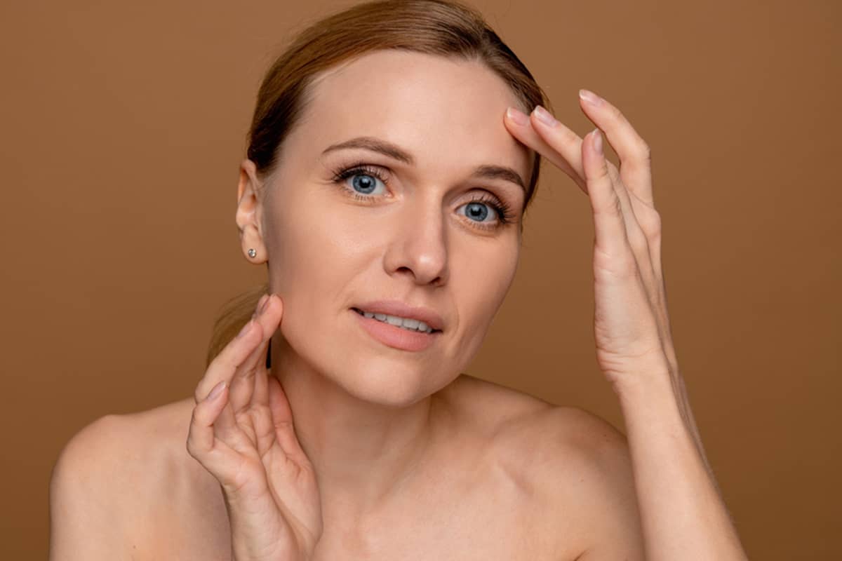 A woman touching her face and jawline to assess skin laxity and structural volume, illustrating the first step in choosing between a thread lift and a deep plane facelift.