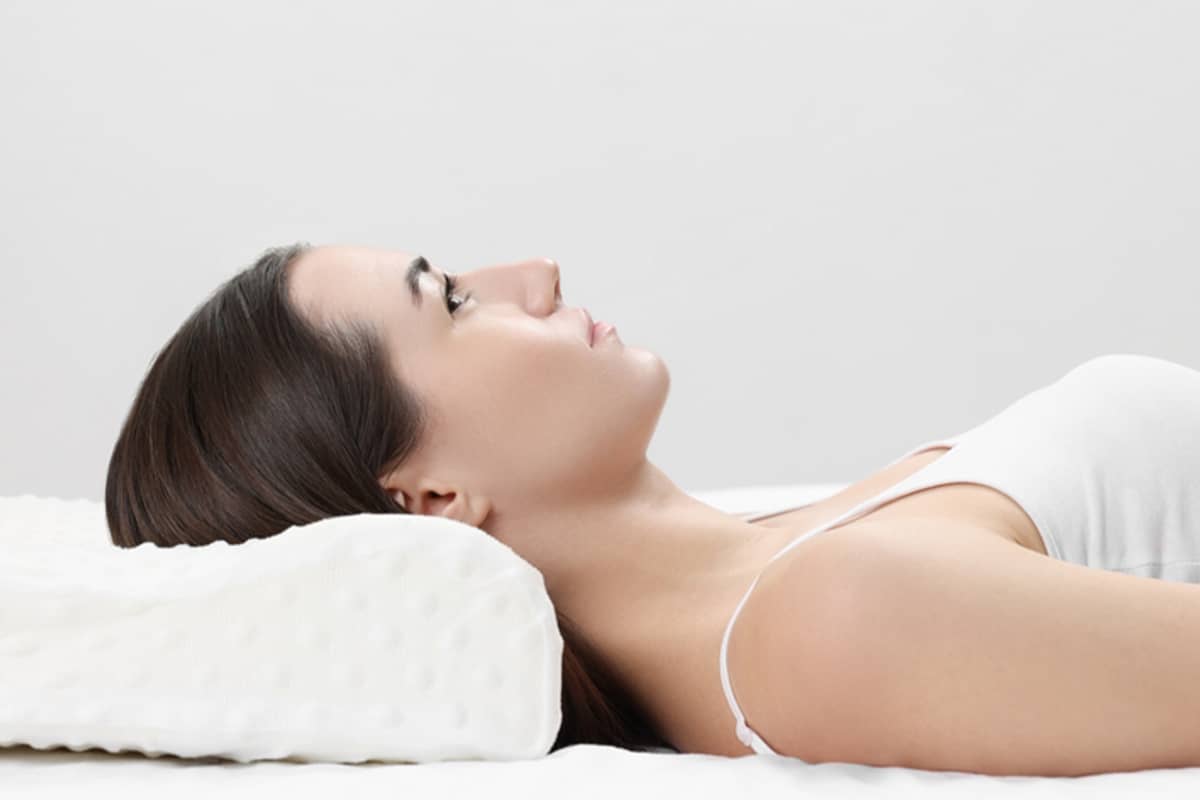 A woman lying on her back with her head elevated on a specialized orthopedic pillow to reduce swelling during her deep plane facelift recovery.