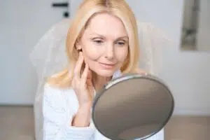 A woman in a white robe looking into a handheld mirror to evaluate her initial facial contours three days after a deep plane facelift.