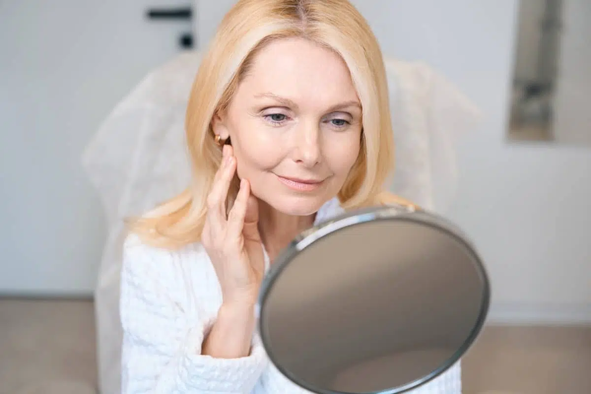 A woman in a white robe looking into a handheld mirror to evaluate her initial facial contours three days after a deep plane facelift.