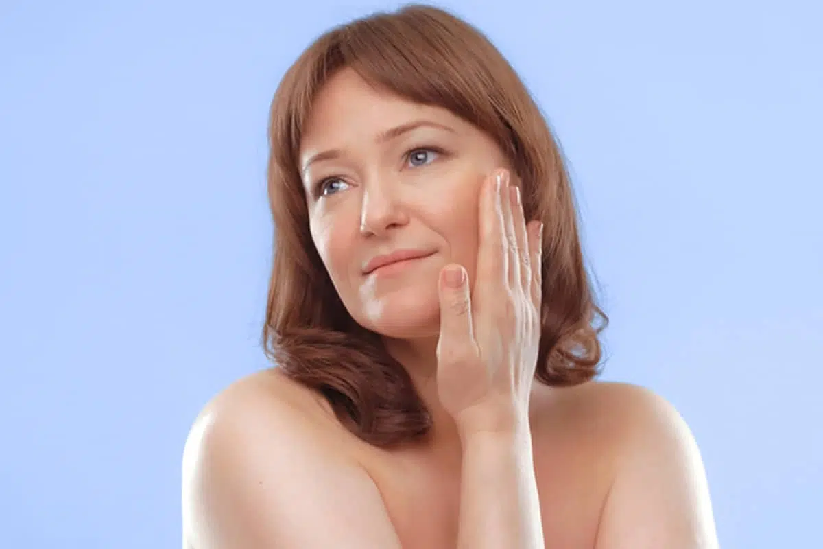 A woman touching the skin near her ear and cheek to identify where deep plane facelift incisions are typically hidden within natural facial contours.