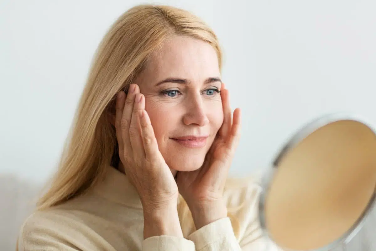 A woman looking in a mirror and touching her temples to monitor the maturation and fading of her facelift scars during the recovery phase.