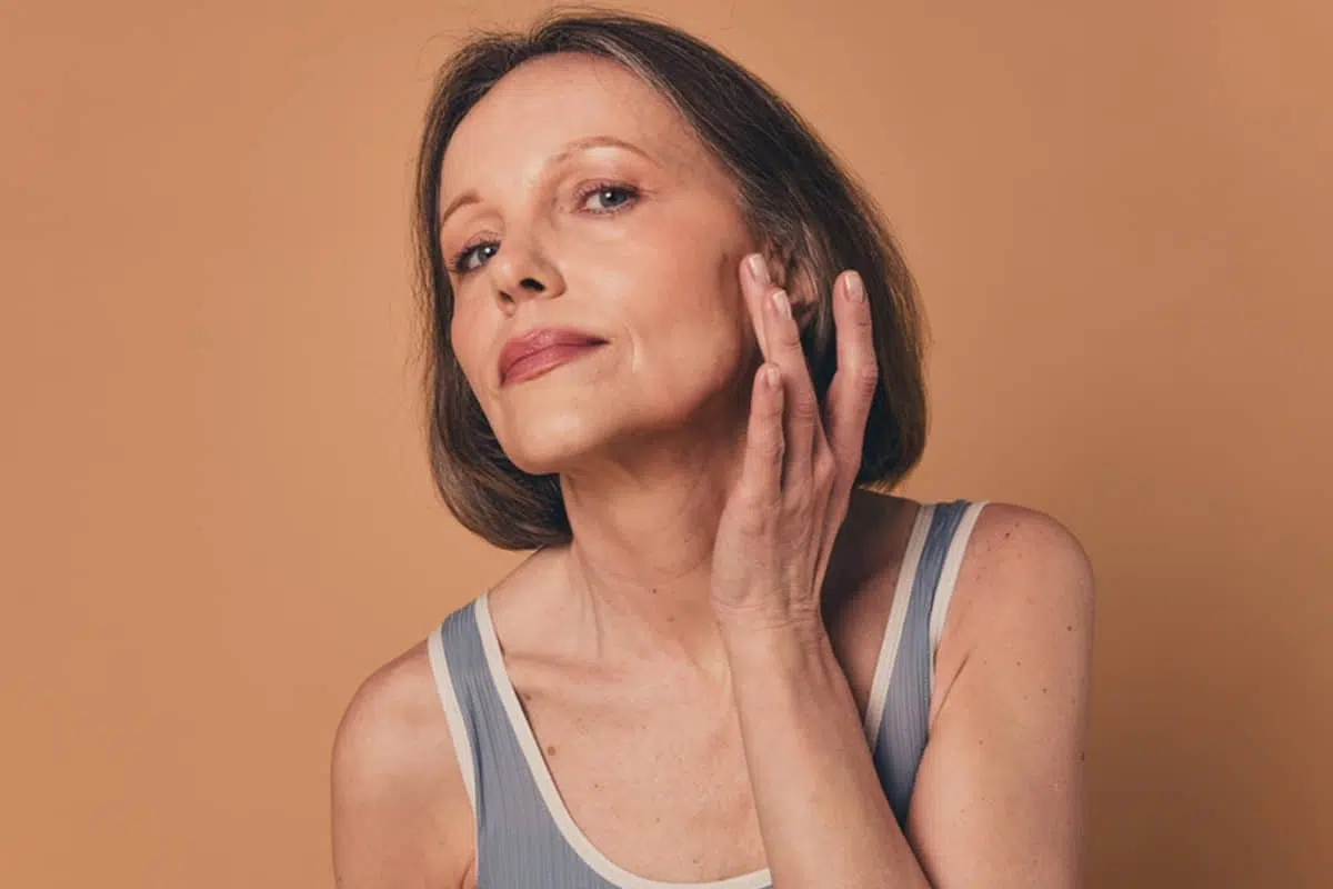 A woman gently touching her cheek and jawline while looking up, assessing her skin’s elasticity before undergoing a facelift.