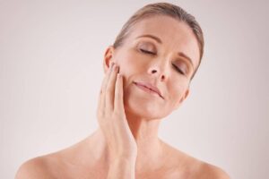 A woman gently touching her cheek and jawline with both hands to evaluate areas of temporary numbness after a deep plane facelift.