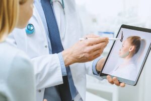 A doctor in a white lab coat uses a stylus to point at a simulated facial lift map on a tablet screen while consulting with a patient.