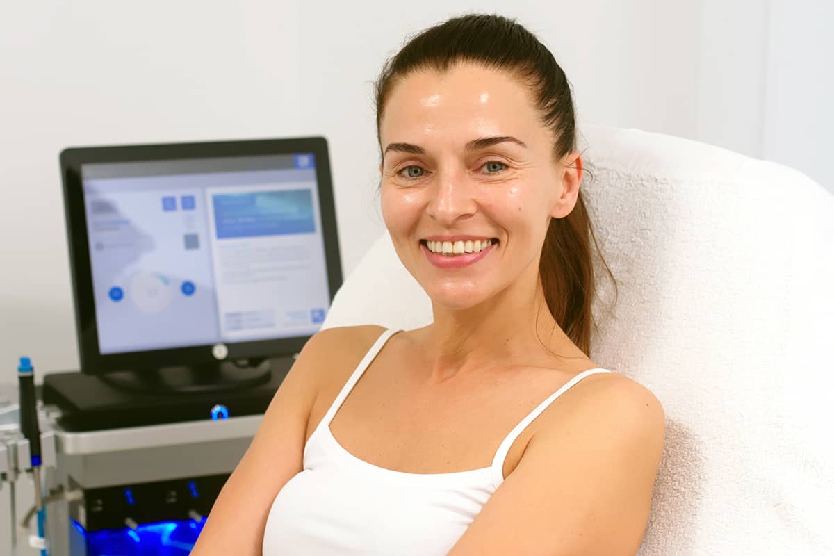 A smiling woman with her hair in a ponytail sits back in a clinical treatment chair with a medical imaging monitor visible in the background.