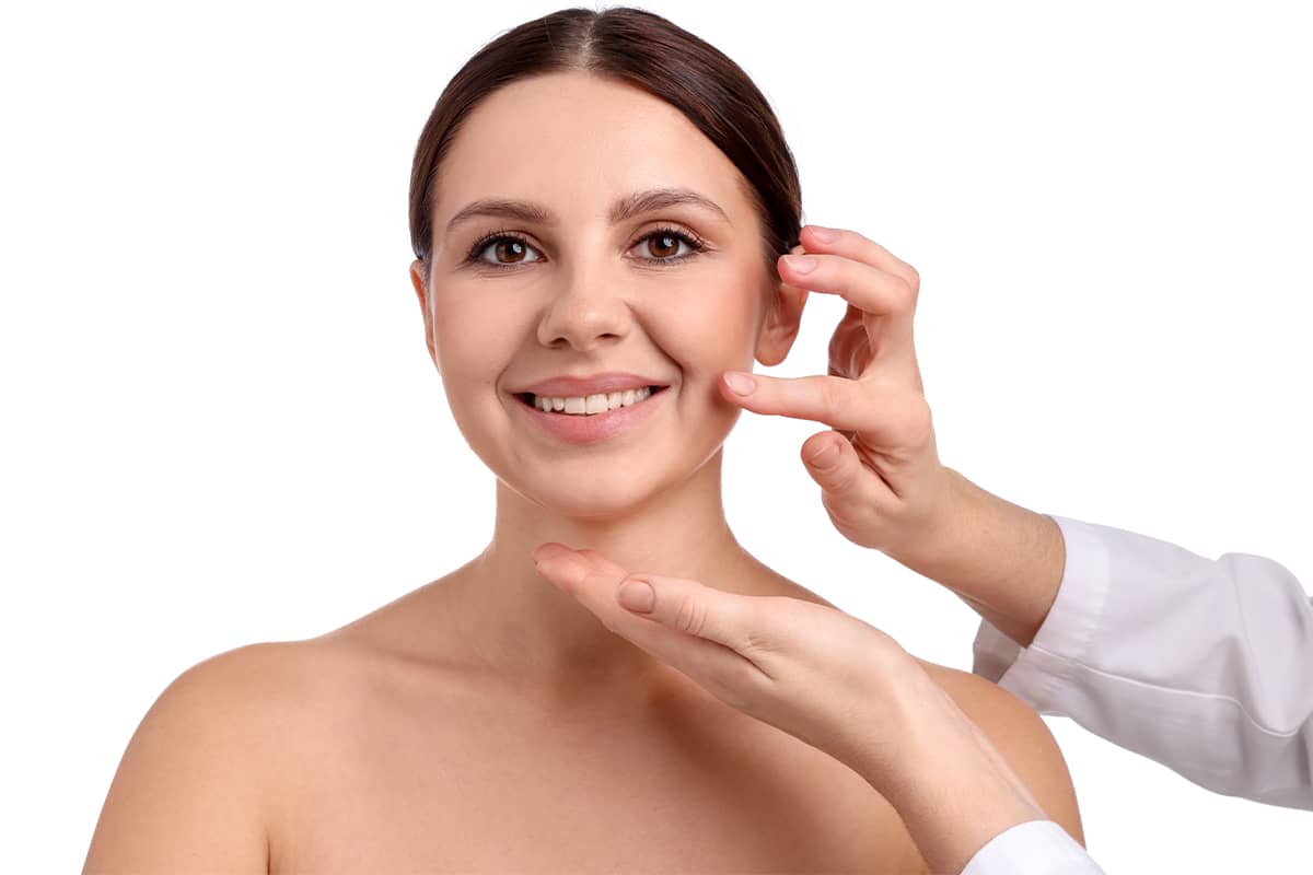 A smiling woman looking forward as a surgeon's hands in a white lab coat gently touch her jawline and check her skin elasticity.