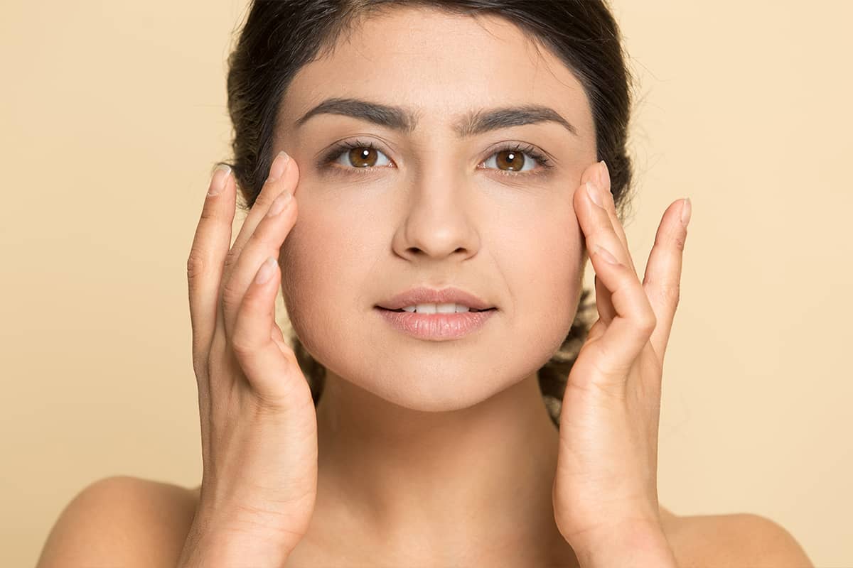 A close-up portrait of a woman with dark hair and brown eyes gently placing her fingertips on her cheeks against a beige background.